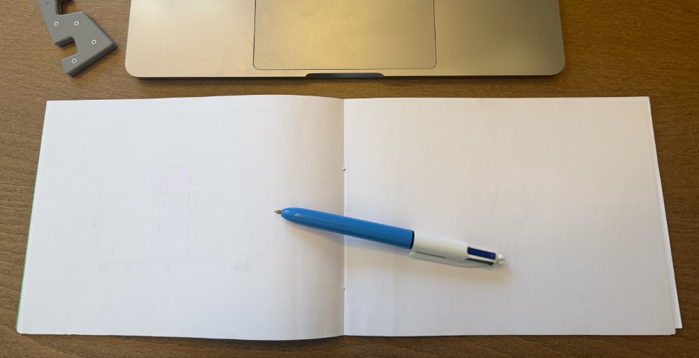 Photo of an A5 notebook opened long-ways underneath a computer laptop keyboard.